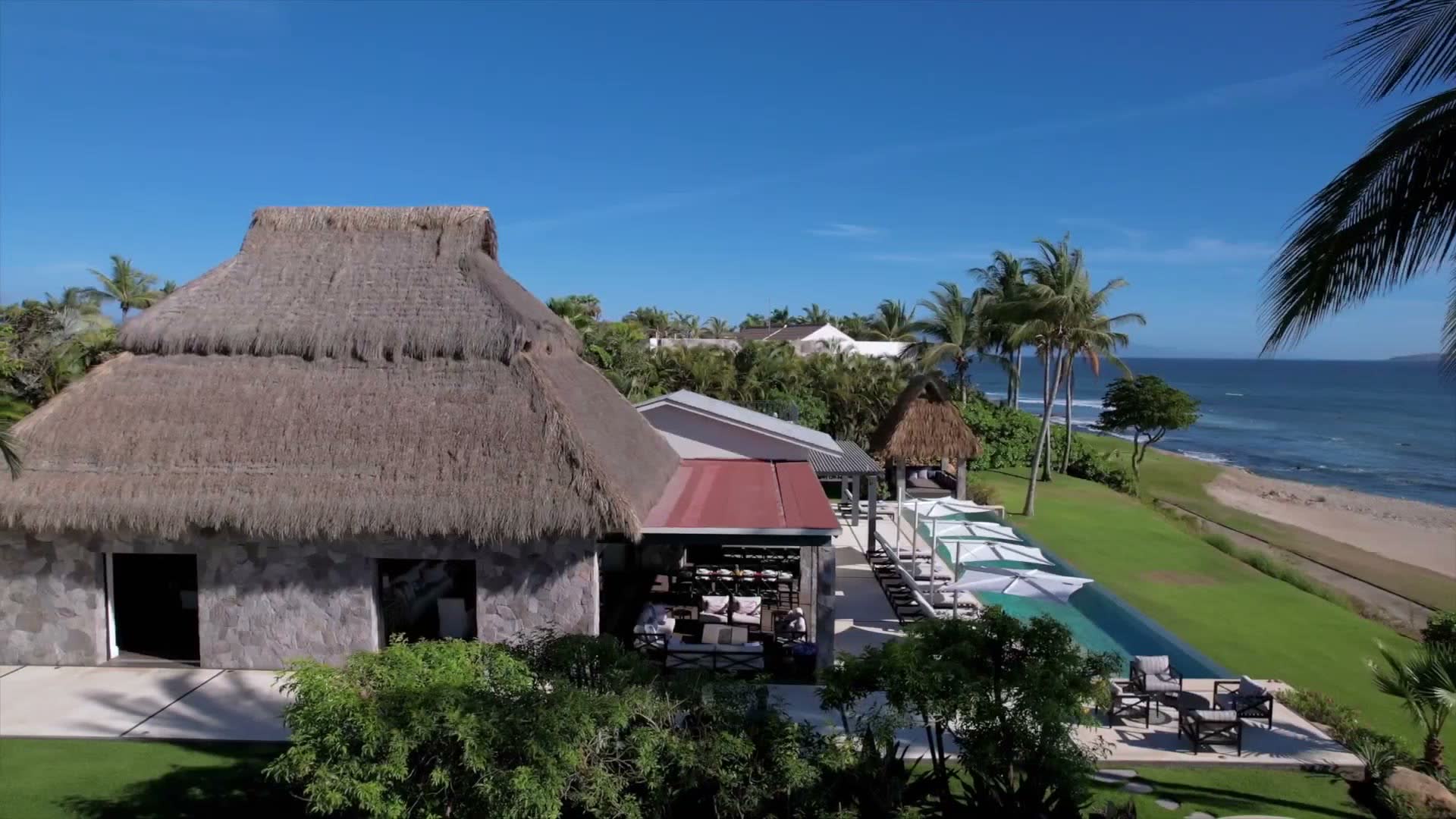 Aerial view of a palapa-roofed beachfront villa with a long infinity pool above the sand.