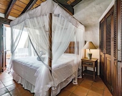 Cozy bedroom at Casa Cosmos, Mexico: four-poster bed, sheer canopy, wooden furnishings, and natural light through open shutters.