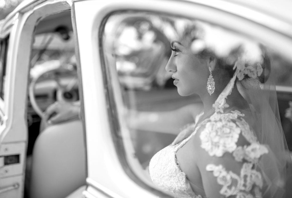 Bride in lace wedding dress sits thoughtfully in vintage car; Los Cabos Wedding Villas make moments unforgettable.