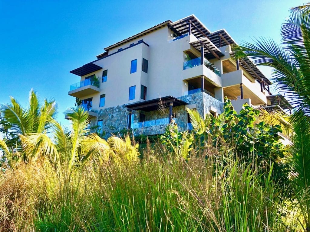 Alt: Modern multi-story villa, Condo Cualli in Mexico, with balconies and lush tropical foliage under a clear blue sky.