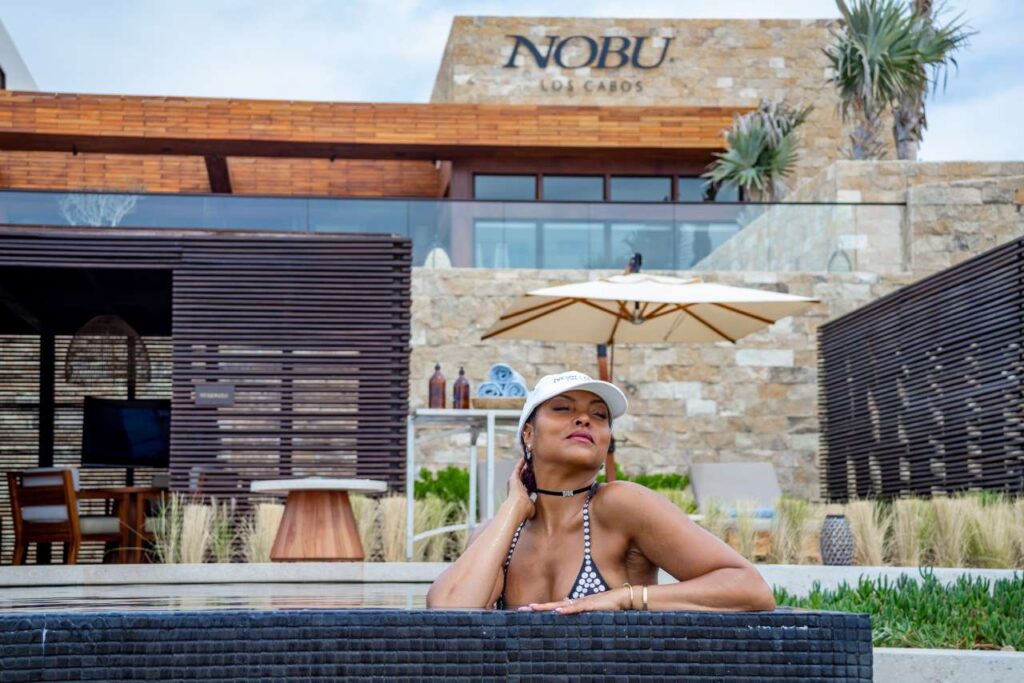Woman in black and white swimsuit by the pool at Nobu Los Cabos, Mexico; elegant brick exterior with desert landscaping.