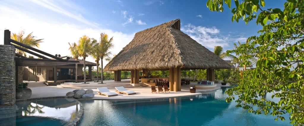 Pool and thatched pavilion beside a modern villa with palm trees