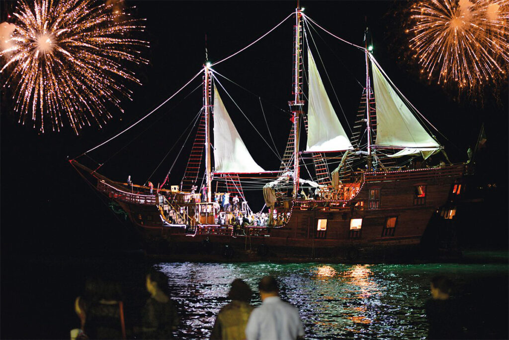 A lit pirate-like ship anchored at night, fireworks overhead, viewers onshore. Luxury vacation villa rental in Mexico.