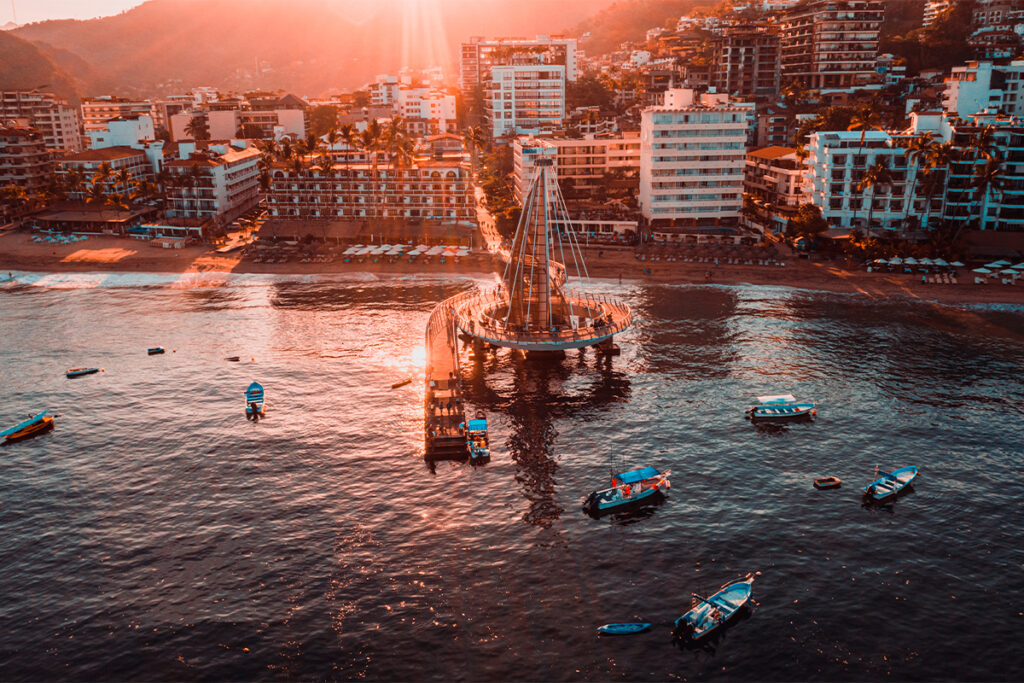 Aerial view of a seaside town in Mexico at sunset with pier, boats, modern buildings, and green hills.