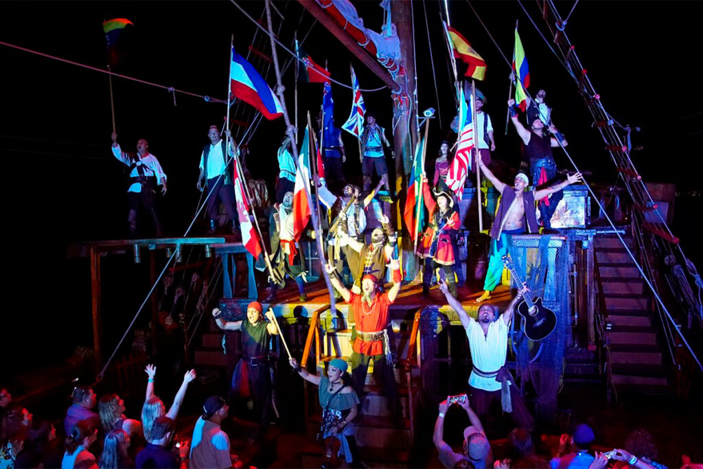 Actors in pirate costumes perform with international flags on a dimly lit ship stage; cheering audience watches.