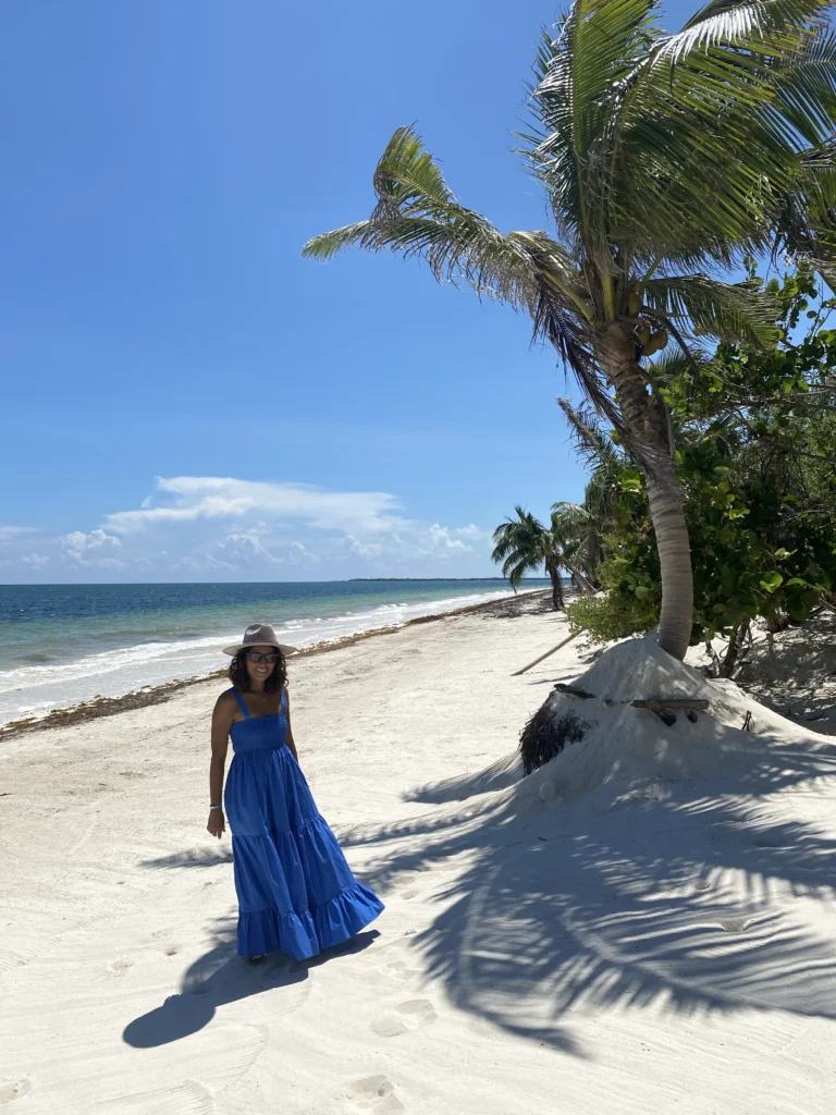 Rocio from Villa Experience, wearing a blue dress and sun hat, walks along a palm-lined beach in Tulum, Mexico, with the ocean and clear blue sky in the background.