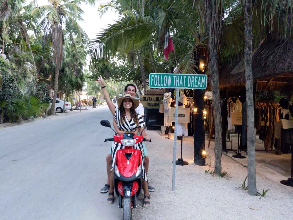 Two people sit on a red scooter on a tropical road lined with palm trees, capturing the essence of luxurious outdoor spaces. The person in front, wearing a striped shirt and straw hat, holds up a peace sign beside a green sign that reads "FOLLOW THAT DREAM." Shops and lights can be seen in the background, enhancing the vibrant atmosphere. The scene perfectly mirrors the relaxed yet sophisticated vibe of luxury accommodations, emphasizing comfort, scenic views, and an inviting environment.