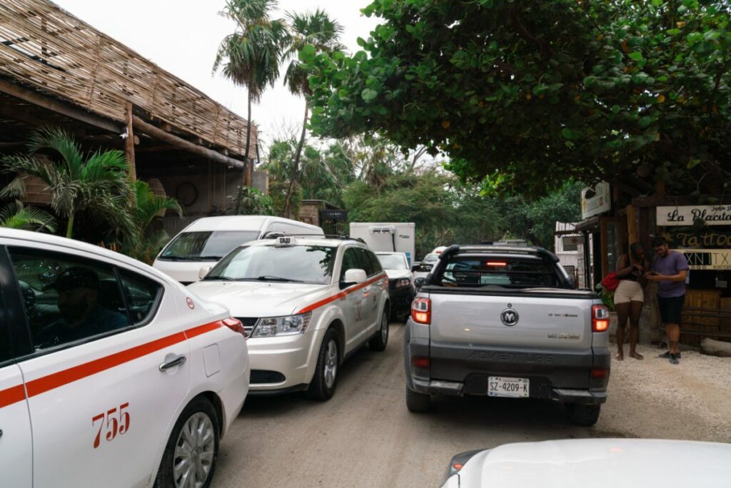A traffic jam in Tulum, Mexico, with white taxis and other vehicles on a narrow street, lined with palm trees and small shops.