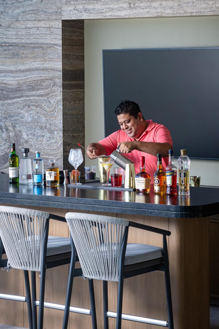 A person in a pink shirt makes a cocktail at Punta Pacifico bar, Punta Mita, Mexico. Sleek counter, stone walls.