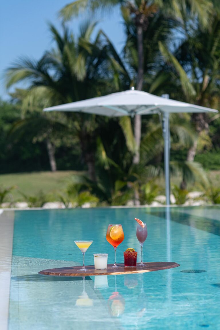 Five colorful cocktails float on a pool tray at Punta Pacifico, Punta Mita, surrounded by lush palms.