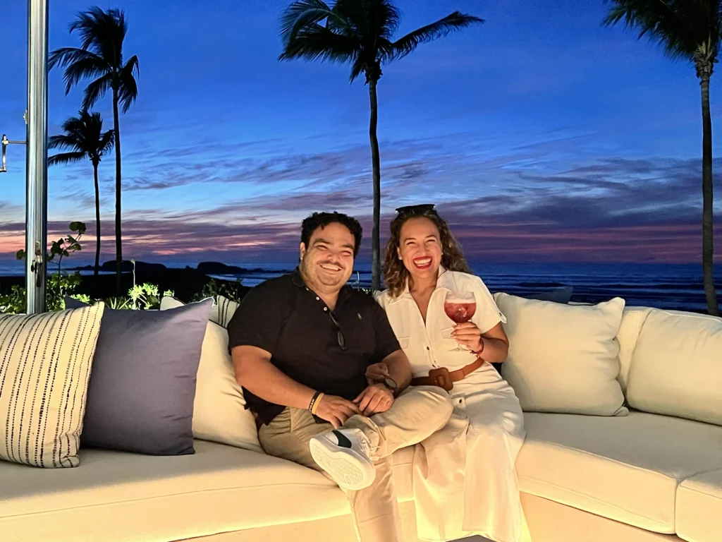 Albriet and Angel from Villa Experience Team relaxes at sunset on a Pacifico Estate couch, Punta Mita, surrounded by palm trees and sea views.